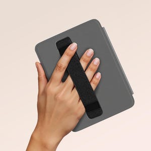 Op de afbeelding: Een grijze tablethoes met een zwarte handriem. De hoes wordt in een hand gehouden, waarbij het ontwerp van de riem wordt getoond. De achtergrond is een zachte, neutrale kleur.