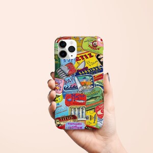 Może przedstawiać: Etui na telefon z kolażem etykiet po puszkach sardynek w stylu vintage w r&oacute;żnych kolorach. Etykiety zawierają tekst i ilustracje, tworząc estetykę retro. Etui jest trzymane w dłoni.