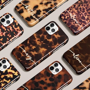 Op de afbeelding: Een verzameling gepersonaliseerde telefoonhoesjes met een schildpadpatroon. De hoesjes hebben een glanzende afwerking en een witte scriptnaam. De kleuren zijn bruin, beige en zwart.