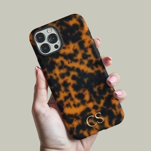 May include: A tortoiseshell-patterned phone case with a black and amber design. The case has a gold-colored monogram of the letters "CS" and a camera cutout. The phone case is held in a hand.