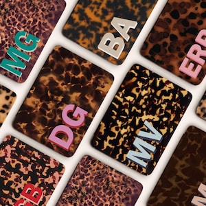 May include: A selection of rectangular cases with a tortoiseshell design. Each case displays a different set of capital letters in various colours, including teal, pink, and white. The cases are arranged on a white surface.