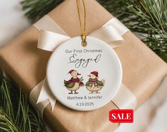 Könnte beinhalten: Weißer Keramik-Weihnachtsschmuck mit goldenem Faden, mit zwei Enten-Illustrationen mit Weihnachtsmützen und Schals. Der Text lautet "Our First Christmas Engaged", Namen und ein Datum. Der Schmuck liegt auf einem Geschenk.