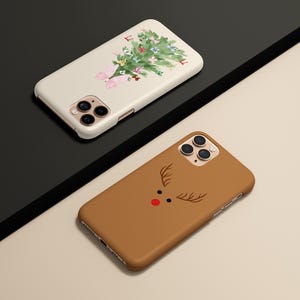 May include: Two phone cases. One is off-white with a watercolor Christmas tree design. The other is brown with a reindeer face design. Both cases have a glossy finish and are angled on a black and beige surface.