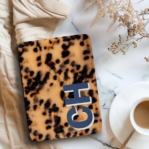 Op de afbeelding: Een rechthoekige hoes met een schildpadpatroon en de letters "CH" in blauw en wit. De hoes ligt op een marmeren oppervlak naast een kopje koffie en een takje gedroogde bloemen. Op de achtergrond is een beige kledingstuk te zien.