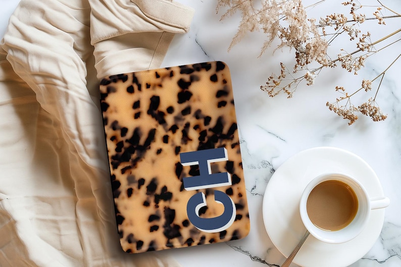 Op de afbeelding: Een rechthoekige hoes met een schildpadpatroon en de letters "CH" in blauw en wit. De hoes ligt op een marmeren oppervlak naast een kopje koffie en een takje gedroogde bloemen. Op de achtergrond is een beige kledingstuk te zien.