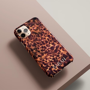 May include: A smartphone case with a leopard print design in shades of brown and tan. The name "SARAH" is printed in white at the bottom. The case is on a neutral-toned surface.