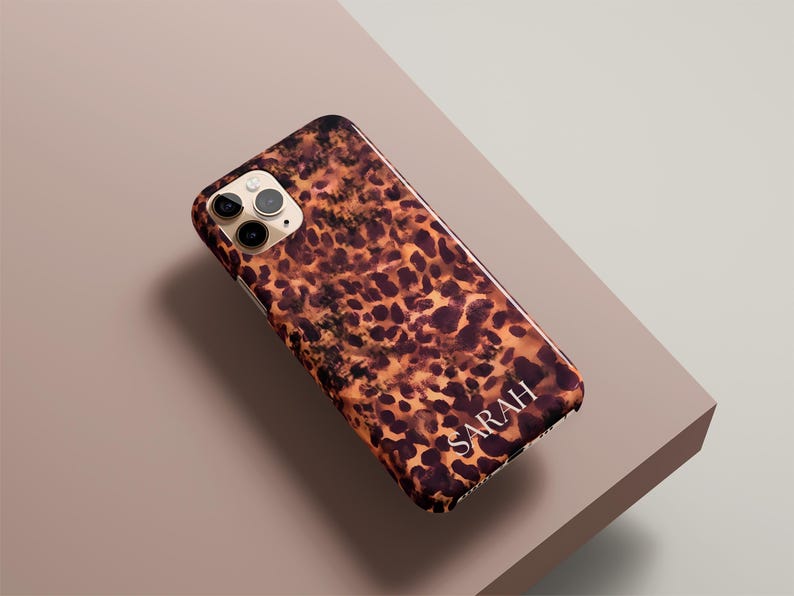 May include: A smartphone case with a leopard print design in shades of brown and tan. The name "SARAH" is printed in white at the bottom. The case is on a neutral-toned surface.