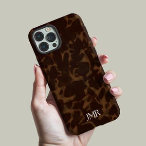 May include: A brown and tan tortoiseshell-patterned phone case with the initials "JMR" in white. The case is held in a hand, showcasing the camera and phone design.