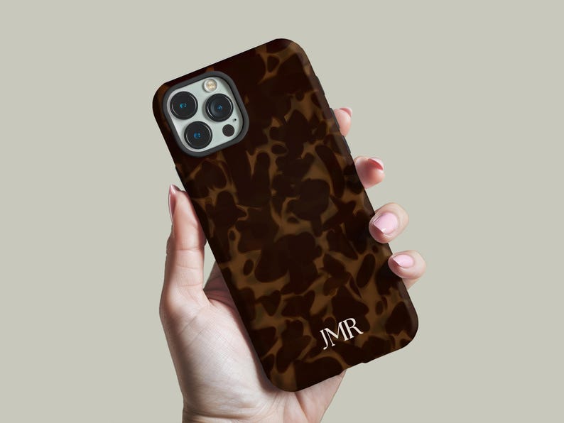 May include: A brown and tan tortoiseshell-patterned phone case with the initials "JMR" in white. The case is held in a hand, showcasing the camera and phone design.