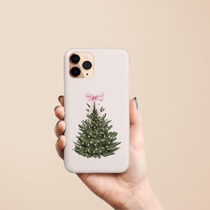 May include: A white phone case featuring a watercolor Christmas tree design. The tree is green with small white lights and a pink bow at the top. The case is held in a hand against a neutral background.