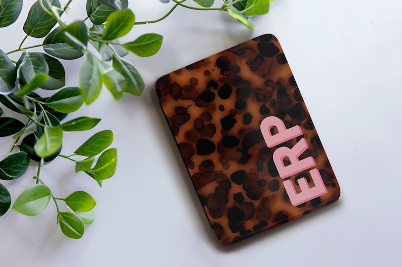 Op de afbeelding: Een rechthoekige telefoonhoes met een schildpadpatroon in bruin- en zwarttinten. De letters "ERP" zijn roze, verticaal op de hoes gestapeld. Groene bladeren bevinden zich in de linkerbovenhoek.