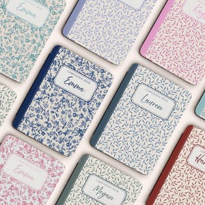 Puede incluir: Cuadernos de composición con estampados florales y de hojas en varios colores, como azul, rosa y verde. Cada cuaderno tiene una etiqueta con un nombre, como "Emma" o "Lauren". Los cuadernos tienen lomos de diferentes colores.