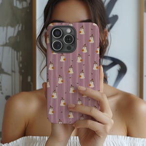 May include: A phone case with a Halloween theme. The case has a pattern of white ghosts wearing witch hats and holding pumpkins, set against a background of vertical purple stripes. The phone case is being held up in front of a person's face.