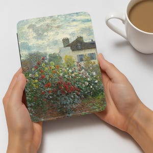 Może przedstawiać: Etui na tablet z kolorowym impresjonistycznym obrazem domu z ogrodem czerwonych, ż&oacute;łtych i białych kwiat&oacute;w. Etui trzymane jest w dw&oacute;ch dłoniach, z białym kubkiem kawy w tle.
