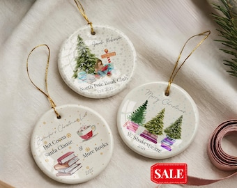 May include: Three round ceramic Christmas ornaments with gold string hangers. Each ornament features a unique design: a Christmas tree scene, a list with "Hot Cocoa" and "Santa Clause", and a "Merry Christmas" message. The ornaments have text that reads "North Pole Book Club" and "W. Shakespeare".