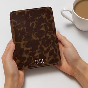 K&ouml;nnte beinhalten: Eine braun-beige gemusterte H&uuml;lle mit den Initialen "JMR" in Wei&szlig;. Die H&uuml;lle wird in zwei H&auml;nden gehalten, mit einer wei&szlig;en Kaffeetasse im Hintergrund. Die H&uuml;lle hat ein Schildpatt-Design.
