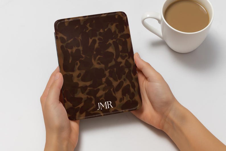 K&ouml;nnte beinhalten: Eine braun-beige gemusterte H&uuml;lle mit den Initialen "JMR" in Wei&szlig;. Die H&uuml;lle wird in zwei H&auml;nden gehalten, mit einer wei&szlig;en Kaffeetasse im Hintergrund. Die H&uuml;lle hat ein Schildpatt-Design.