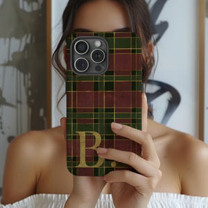 May include: A phone case with a tartan design in burgundy, green, and gold. The case displays the letter "B" in gold. The case has a camera cutout with three lenses.