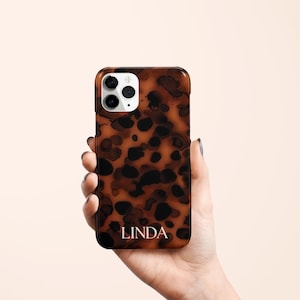 May include: A brown and black tortoiseshell phone case with the name "LINDA" in white lettering. The case has a glossy finish and a camera cutout. The phone case is held in a hand.