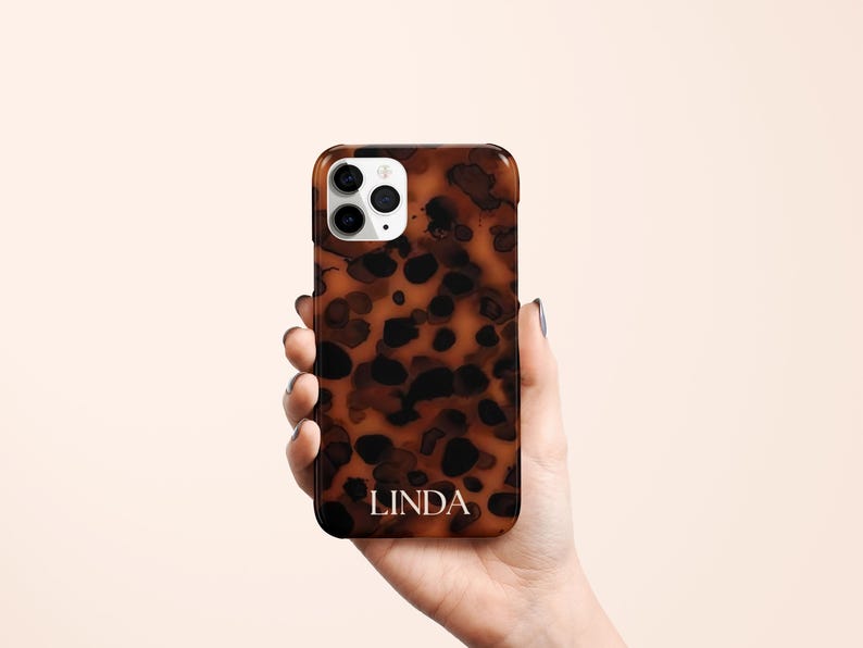 May include: A brown and black tortoiseshell phone case with the name "LINDA" in white lettering. The case has a glossy finish and a camera cutout. The phone case is held in a hand.
