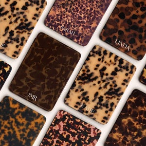 May include: A selection of rectangular cases featuring a tortoiseshell design in shades of brown, tan, and black. Some cases are personalised with names such as "KATE", "SARAH", and "LINDA" in white lettering. The cases are displayed on a white background.