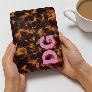 Op de afbeelding: Een bruin en tan patroon hoesje met de letters "DG" in roze. De hoes wordt in twee handen gehouden. Een witte mok met een bruine vloeistof staat op de achtergrond.