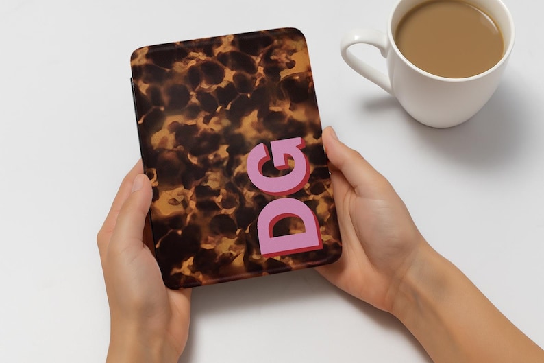 Op de afbeelding: Een bruin en tan patroon hoesje met de letters "DG" in roze. De hoes wordt in twee handen gehouden. Een witte mok met een bruine vloeistof staat op de achtergrond.
