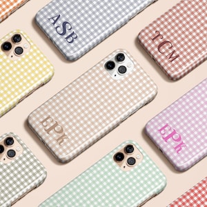May include: A collection of phone cases in various colors, each featuring a gingham pattern. Several cases display initials in a contrasting color. Colors include yellow, gray, pink, beige, green, and red. The cases are arranged on a light pink surface.