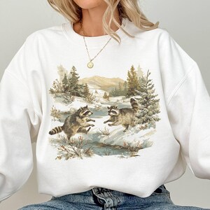May include: White sweatshirt featuring a watercolor illustration of two raccoons playing in a snowy landscape with trees and mountains in the background. The sweatshirt has long sleeves and a crew neck.