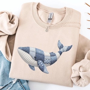 May include: A beige sweatshirt featuring a blue whale design. The whale is made of patchwork fabric in various shades of blue. The sweatshirt is folded, with a pair of blue jeans visible in the background.
