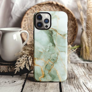 May include: A smartphone case with a marble design in shades of green and gold. The case has a black border and a camera cutout. The background includes a white pitcher, dried plants, and a woven basket.