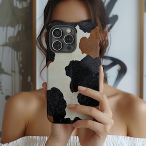 May include: A smartphone case with a cowhide pattern. The case features a mix of black, brown, and white patches, creating a natural, textured look. The case has a black border and cutouts for the phone's camera lenses.