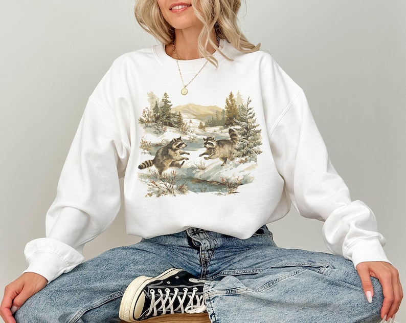 May include: White sweatshirt featuring a watercolor illustration of two raccoons playing in a snowy landscape with trees and mountains in the background. The sweatshirt has long sleeves and a crew neck.