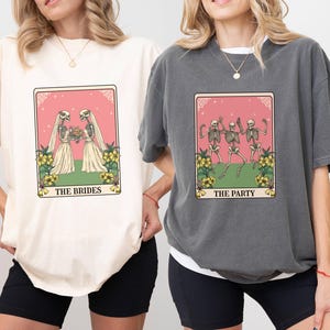 May include: Two oversized t-shirts with a skeleton themed graphic. The ivory shirt features two skeletons getting married with the text "The Brides". The gray shirt features three skeletons dancing with the text "The Party".