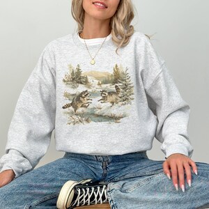 May include: A light gray sweatshirt featuring a nature scene with two raccoons playing in a snowy landscape. The graphic includes trees, a stream, and mountains in the background. The sweatshirt is a crew neck style.