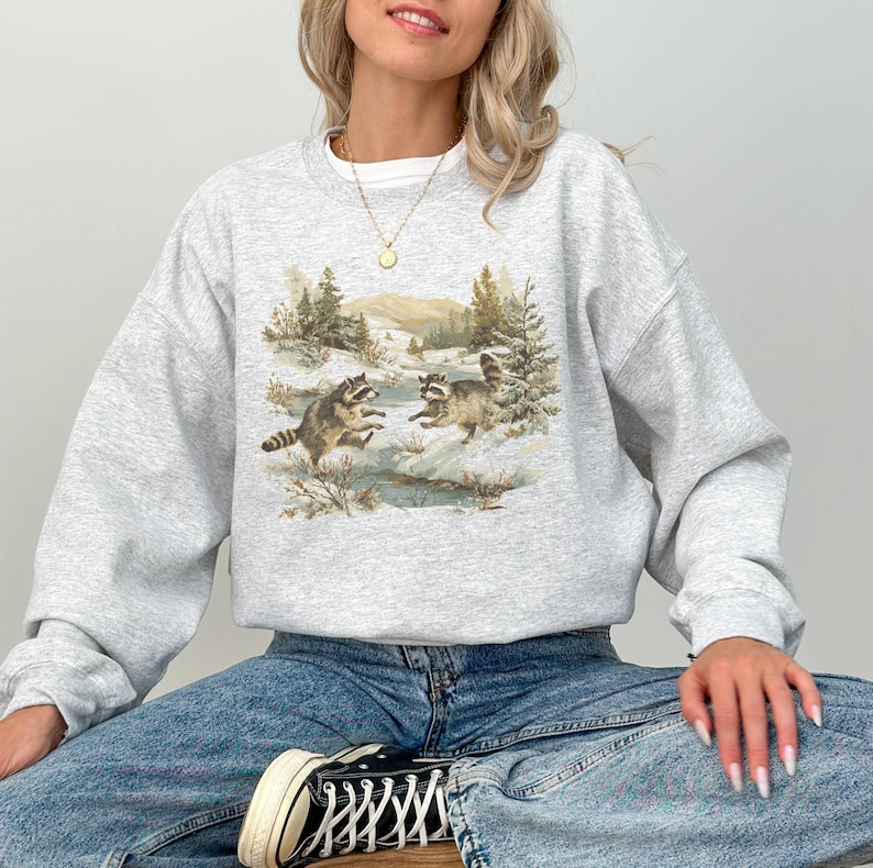 May include: A light gray sweatshirt featuring a nature scene with two raccoons playing in a snowy landscape. The graphic includes trees, a stream, and mountains in the background. The sweatshirt is a crew neck style.