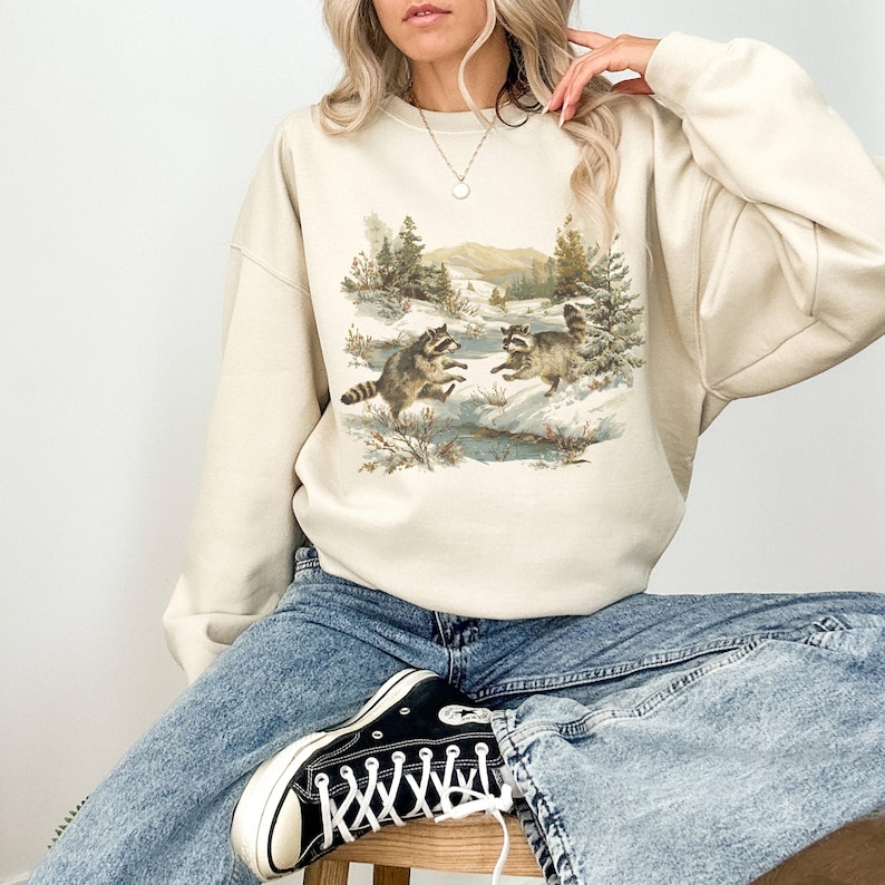May include: Beige sweatshirt featuring a nature scene with two raccoons in a snowy landscape. The graphic includes trees, mountains, and a stream. The sweatshirt is paired with blue jeans and black and white sneakers.
