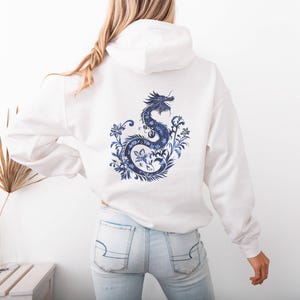 May include: White hoodie with a blue and white dragon design on the back. The dragon is surrounded by floral details.