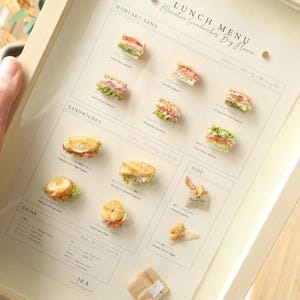 以下が含まれることがあります： ミニチュアサンドイッチとサイドメニューが掲載された、額入りの「LUNCH MENU」。メニューには「Wampaku Sand」と「Sandwiches」のセクションがあり、各アイテムの詳細なイラストが掲載されています。フレームは明るい色の木材で、メニューはクリーム色の紙に印刷されています。