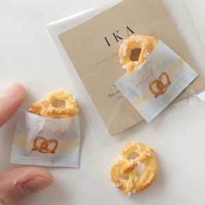 Op de afbeelding: Miniatuur pretzels met grove zoutkristallen, gepresenteerd in kleine papieren zakjes. De zakjes zijn voorzien van het opschrift "pretzel day" met een pretzel-afbeelding. Op de achtergrond is een bruine kaart met de tekst "IKA" te zien.