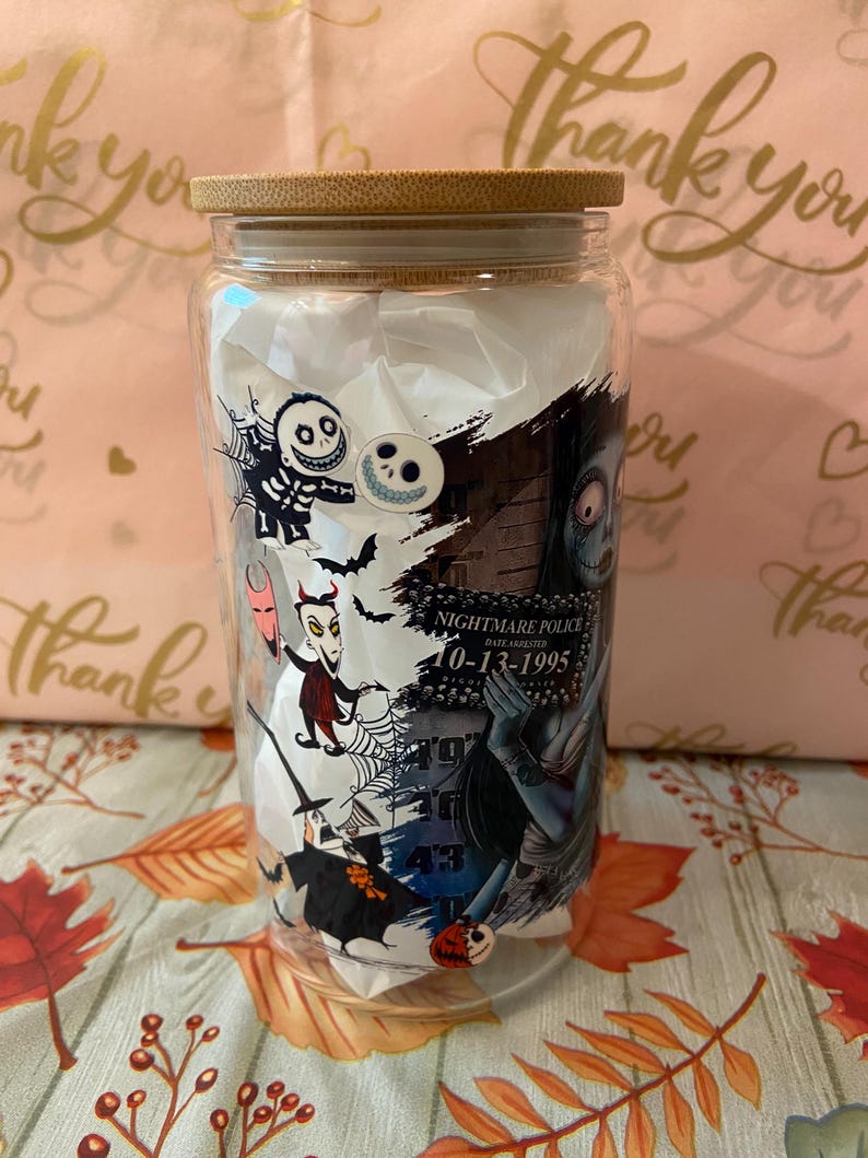 May include: A clear glass tumbler with a bamboo lid features a colorful graphic print of characters from the Nightmare Before Christmas movie. The print includes Jack Skellington, Sally, Oogie Boogie, and other characters. The text "Nightmare Police" and "10-13-1995" is visible on the print.