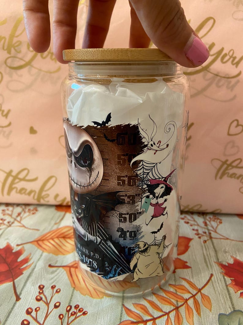 May include: A glass jar with a bamboo lid featuring a Nightmare Before Christmas design. The jar has a black and white image of Jack Skellington, Sally, and Zero. The jar also has a measurement scale on the side.
