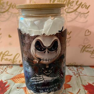 May include: A glass jar with a bamboo lid featuring a graphic of Jack Skellington from The Nightmare Before Christmas. The graphic shows Jack Skellington smoking a cigar and holding a cat. The text "Nightmare P.D. 10-25-1978" is printed on the jar.