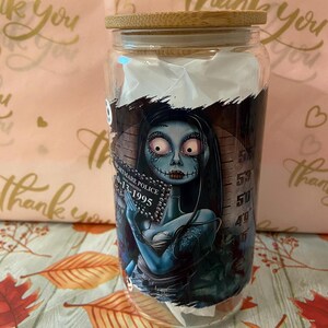 May include: A glass jar with a bamboo lid featuring a graphic of a blue-skinned woman with black hair and a black dress. The woman is holding a book with the text "Nightmare Police 10-13-1995". The jar has a white background with a black and white striped pattern.