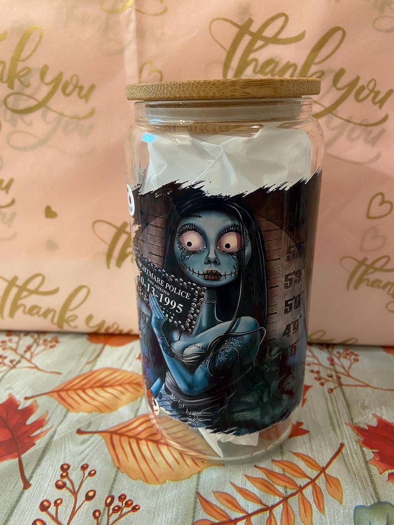 May include: A glass jar with a bamboo lid featuring a graphic of a blue-skinned woman with black hair and a black dress. The woman is holding a book with the text "Nightmare Police 10-13-1995". The jar has a white background with a black and white striped pattern.
