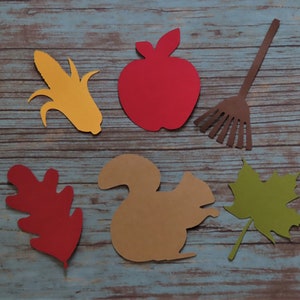 May include: Paper cutouts of autumnal objects, including a red apple, a yellow corn cob, a brown rake, a red oak leaf, a brown squirrel, and a green maple leaf.
