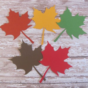 May include: Five paper maple leaves in different colors: red, orange, yellow, green, and brown. The leaves are arranged in a circle on a whitewashed wooden background.