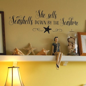 May include: A white shelf with a seashell quote that reads "She sells seashells down by the seashore" with a black starfish in the middle. There are seashells and starfish on the shelf, and a doll wearing a black and white polka dot dress is sitting on the edge of the shelf.