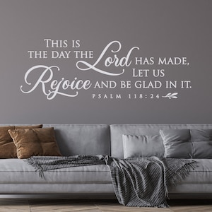 May include: White vinyl wall decal with the text "This is the day the Lord has made, Rejoice and be glad in it. Psalm 118:24" on a gray wall above a gray couch with brown and white throw pillows and a gray throw blanket.