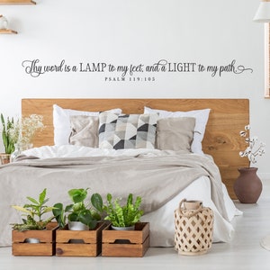 May include: A bedroom with a wooden headboard and a beige duvet. The wall has a black vinyl decal with the text "Thy word is a LAMP to my feet, and a LIGHT to my path. PSALM 119:105". There are three wooden crates with potted plants and a woven lantern on the floor.